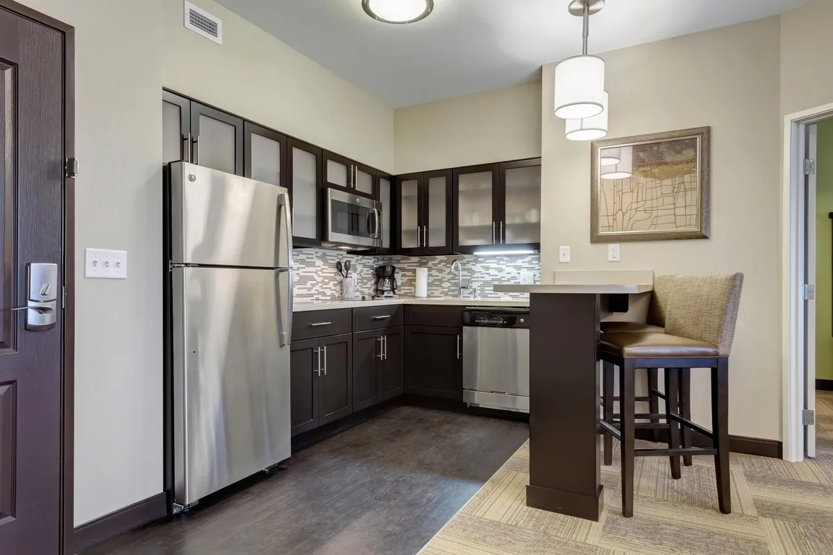 A modern kitchen featuring a stainless steel refrigerator, dark cabinetry, bar seating area, and stylish light fixtures. The space is designed for both cooking and entertaining, with ample room for meal preparation and casual dining. Decorative wall art adds a welcoming touch.