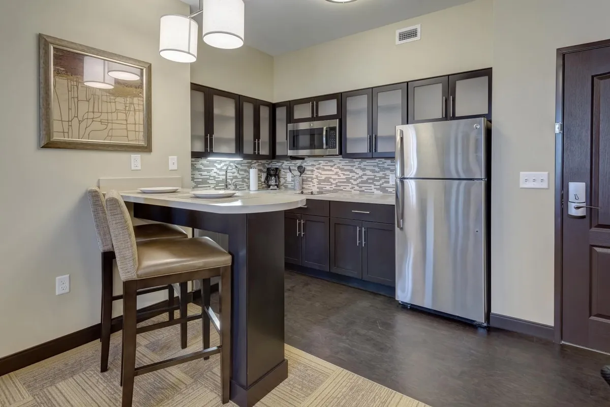 A contemporary kitchen setting with a prominent kitchen island, stainless steel refrigerator, pendant lights, and decorative wall art. The open layout makes it ideal for cooking, sharing meals, or enjoying a cup of coffee.