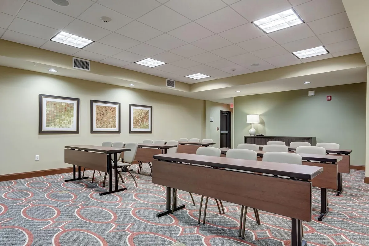 Rows of tables and chairs fill a stylish common area adorned with framed artwork and softly lit by side lamps. The layout suggests a flexible space for dining, working, or socializing, reflecting the multi-use comfort that guests enjoy. The décor is warm and contemporary, with an inviting ambiance.
