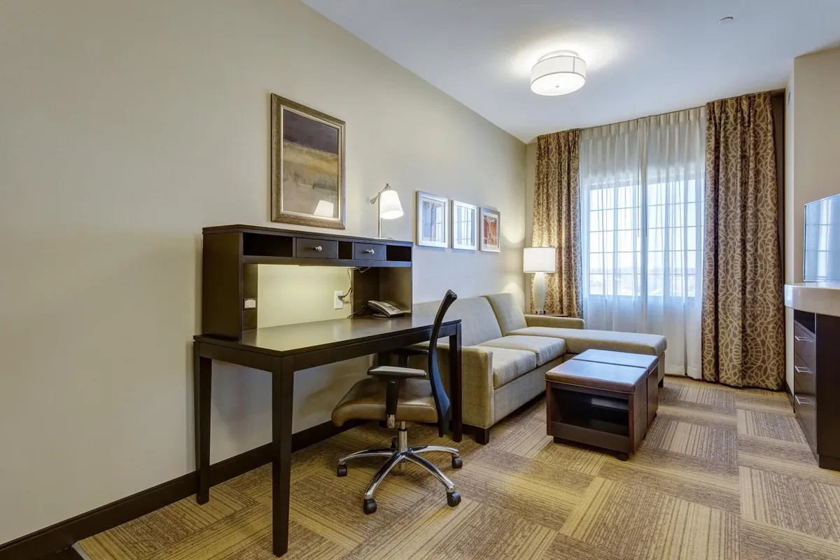 The image captures a bright and inviting interior with a dedicated work desk and chair, a comfortable sofa perfect for relaxation, a window allowing soft morning light to fill the room, and a warm lamp creating a cozy ambiance. The arrangement suggests a perfect blend of comfort and functionality, ideal for both productivity and unwinding after a day exploring St. Louis.