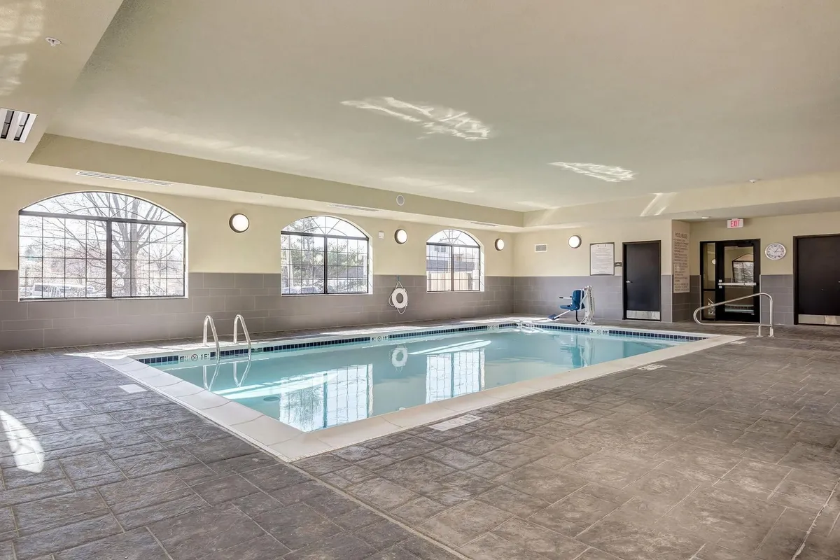 A spacious, sunlit indoor swimming pool area featuring large windows that flood the space with natural light. Poolside seating is available for guests to relax, and safety equipment is visible, reinforcing comfort and family-friendly appeal. There are guests swimming and socializing, emphasizing both recreation and relaxation.