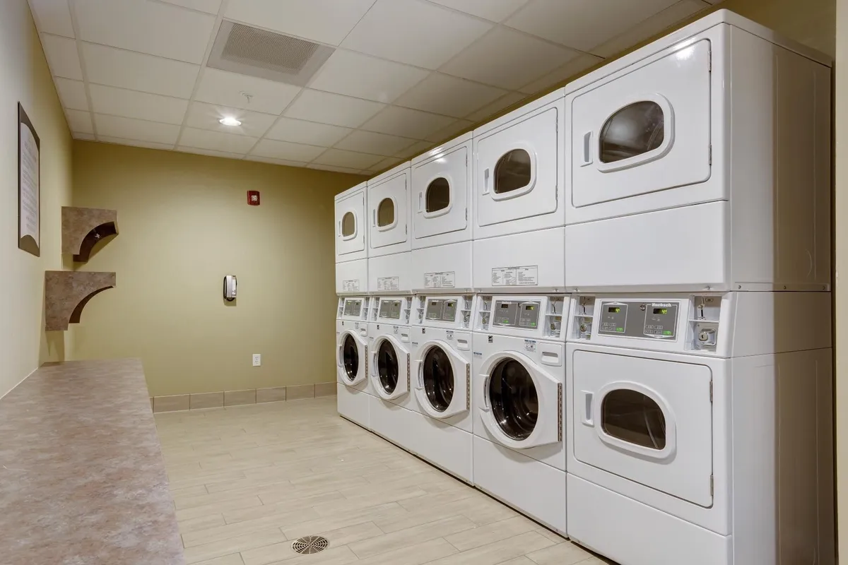 A tidy laundry area with a row of modern washing machines, a practical countertop for folding clothes, and wall-mounted shelves for supplies. The room is organized and well-lit, conveying an atmosphere of efficiency and cleanliness. Guests are seen taking care of their laundry, emphasizing self-sufficiency and the ease of maintaining fresh clothes during a stay.