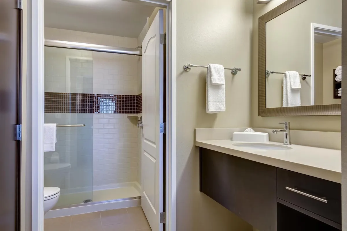 A bright, modern bathroom featuring a glass-enclosed shower area, a vanity with a spacious sink, and a large mirror. The setting is clean, well-lit, and exudes a sense of calm and relaxation—ideal for unwinding after a long day. Soft towels are visible, and the countertop is arranged with complimentary toiletry items, suggesting both practicality and comfort.