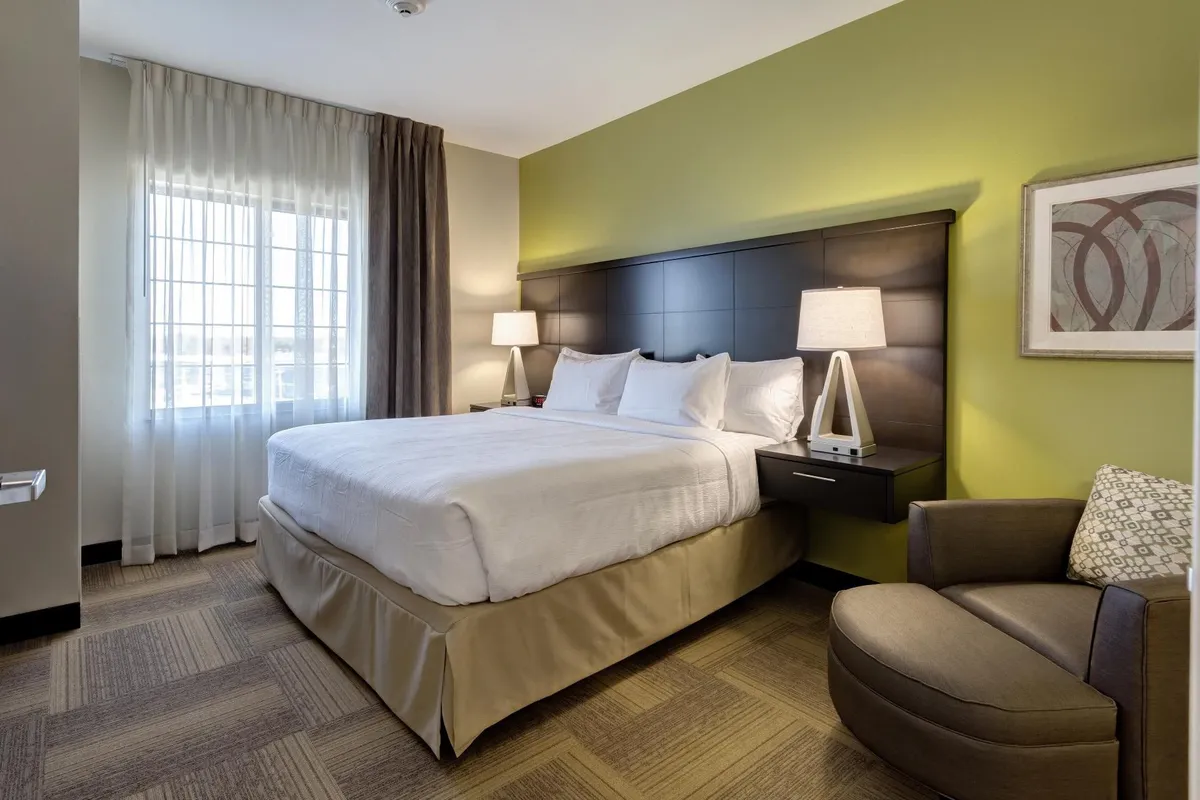 An inviting bedroom with a comfortable bed, an armchair by a bright window, and tasteful decorative artwork. The space exudes warmth and quiet, ideal for reading, relaxing, or enjoying a moment of peace.