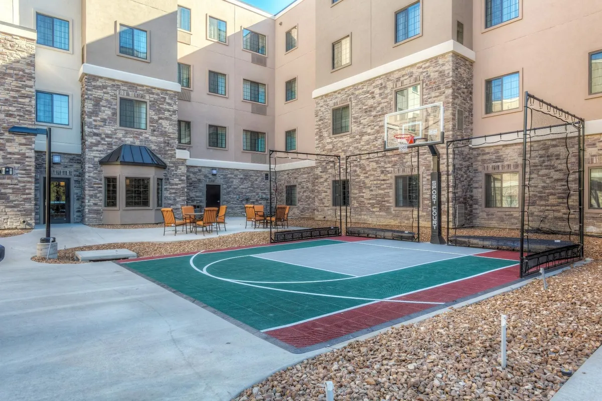 This exterior shot features the Staybridge Suites building with a prominent basketball court and outdoor seating area in the foreground. The architectural details of the hotel are clearly visible, and the sunny setting conveys energy and approachability. The scene is lively and welcoming, suggesting communal activities and spaces for relaxation between work commitments.